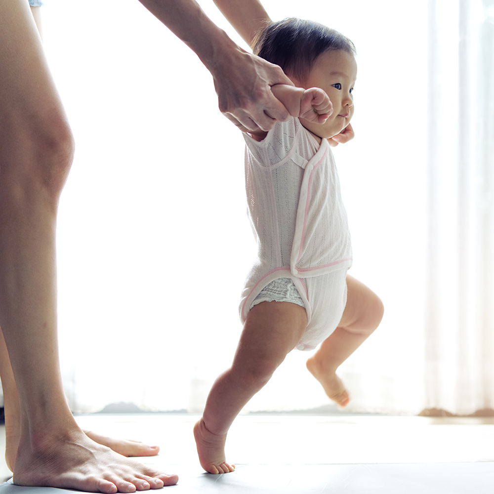 Asian baby taking first steps walk forward on the soft mat. Happy little baby learning to walk with mother help at home. Mother teaching how to walk gently. Baby growth and development concept.