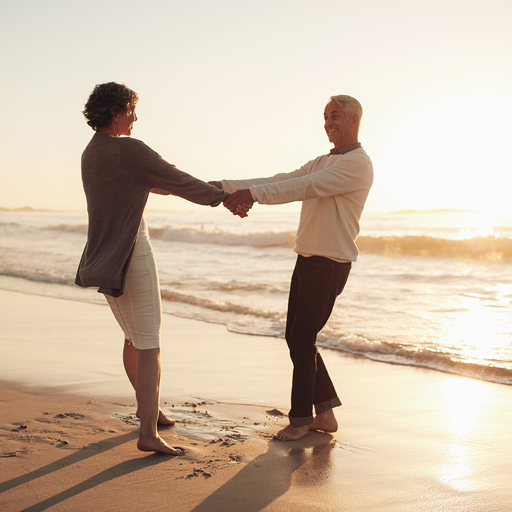 Full length outdoor shot of senior couple holding hands standing on the beach. Mature man and woman playing on the seashore at sunset.